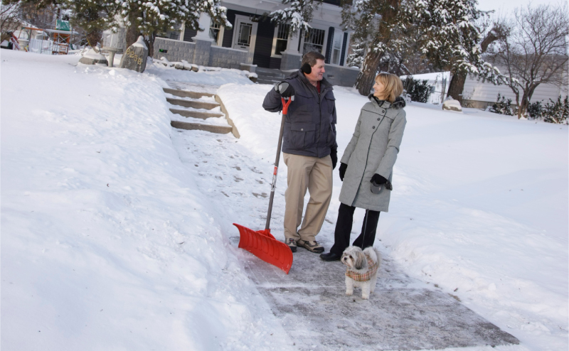 Two people talking on the front sidewalk to a house, one of them is holding a snow shovel.