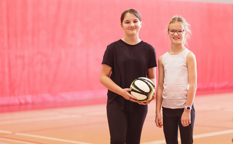 Two girls in a gymnasium. One is holding a volleyball.