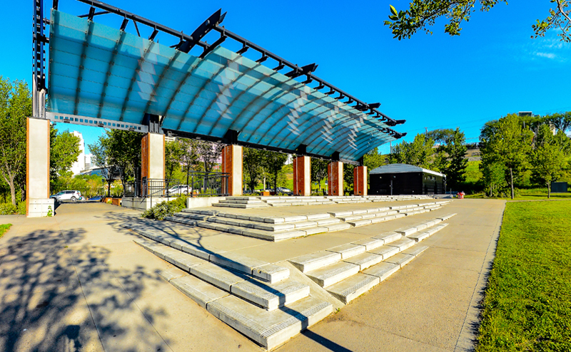 Photo of the Shumka Stage at Louise McKinney Riverfront Park. The stage is a raised platform with steps, covered by a translucent awning overhead.
