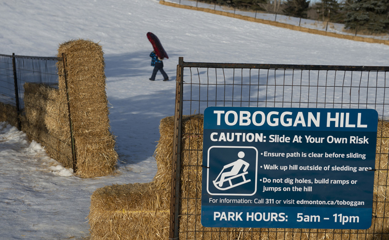 A child walking up a toboggan hill. A sign at the bottom reads "Toboggan Hill. Caution: Slide at your own risk. Ensure path is clear before sliding. Walk up hill outside of sledding area. Do not dig holes, build ramps or jumps on hill. For information, call 311 or visit edmonton.ca/toboggan. Park hours: 5am-11pm"