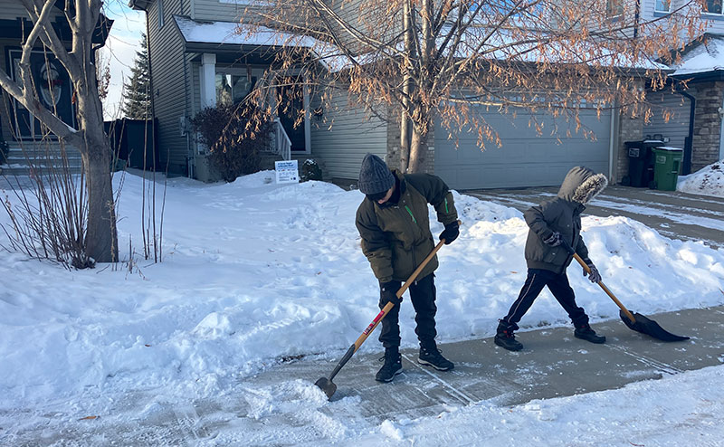 Two teenagers shoveling a residential sidewalk.
